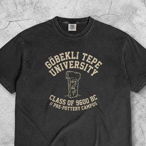 May include: A dark gray t-shirt with the text "GÖBEKLİ TEPE UNIVERSITY" and "CLASS OF 9600 BC // PRE-POTTERY CAMPUS" in a vintage style. The shirt features a graphic of a stone structure. The word "PEPPER" is at the bottom right.