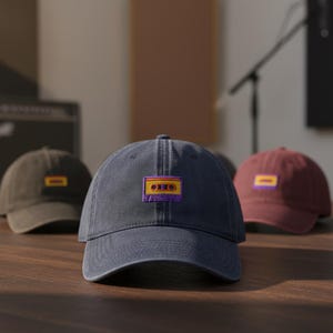 May include: Three baseball caps on a wooden surface. The front cap is navy blue with a colorful cassette tape patch. The other two caps are brown and maroon, each with a rectangular patch. The caps are in a studio setting.