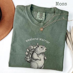 May include: A moss green Comfort Colors t-shirt with the words "weekend warrior" and a graphic of a groundhog wearing a backpack. A silver necklace with a small pendant is on the shirt. A brown hat is in the upper left corner.