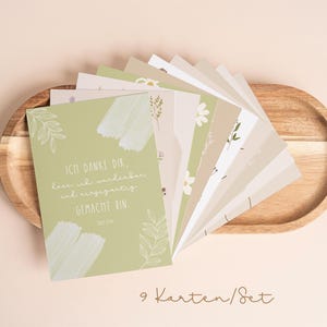 Könnte beinhalten: Ein Set aus neun Karten mit floralen Designs und deutschem Text, angeordnet auf einem Holztablett. Die Karten zeigen eine Vielzahl von Pastellfarben, darunter Grün, Beige und Weiß, mit botanischen Illustrationen.