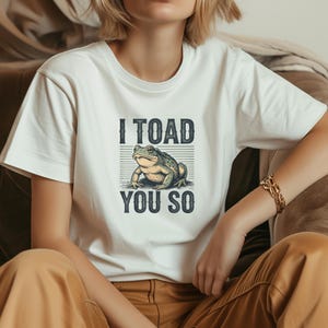 May include: White t-shirt featuring a green and tan toad graphic with the text "I TOAD YOU SO" in dark blue. The toad illustration is in the centre, with the text above and below. The t-shirt has short sleeves.