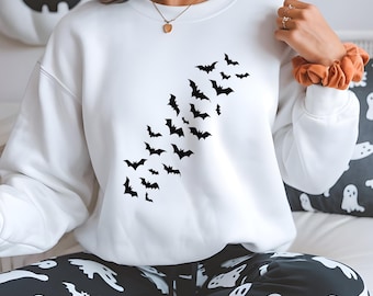 May include: White sweatshirt featuring a diagonal pattern of black bat silhouettes. The long-sleeved top is part of a Halloween-themed ensemble, paired with black and white ghost-patterned pajama pants. A gold necklace and orange scrunchie are also visible.