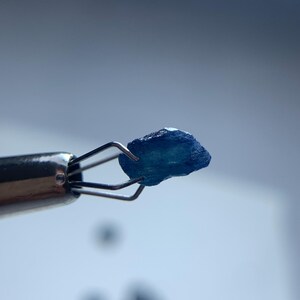 May include: A close-up of a small, rough-cut, blue gemstone held by a silver jeweler's tool. The gemstone has a textured surface and irregular shape. The background is a soft, out-of-focus gradient of white and blue.