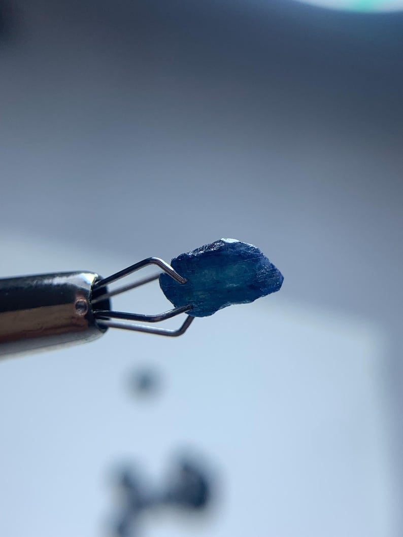 May include: A close-up of a small, rough-cut, blue gemstone held by a silver jeweler's tool. The gemstone has a textured surface and irregular shape. The background is a soft, out-of-focus gradient of white and blue.