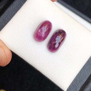 May include: Two oval-shaped, polished ruby gemstones. The rubies are a deep, rich red color with some inclusions visible. They are displayed on a white surface, likely for presentation or sale. The gemstones are a natural, organic shape.