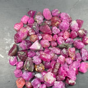 Puede incluir: Una colección de piedras preciosas de rubí en bruto y sin pulir en varios tonos de rosa vibrante y magenta intenso. Las piedras presentan formas irregulares y dentadas con texturas cristalinas naturales, algunas con inclusiones y depósitos minerales de tono metálico.