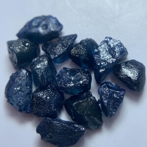 May include: A collection of rough, dark blue gemstones with a textured surface. The stones vary in size and shape, with a deep, saturated color. The gemstones are scattered on a white surface, creating a contrast that highlights their color and texture.