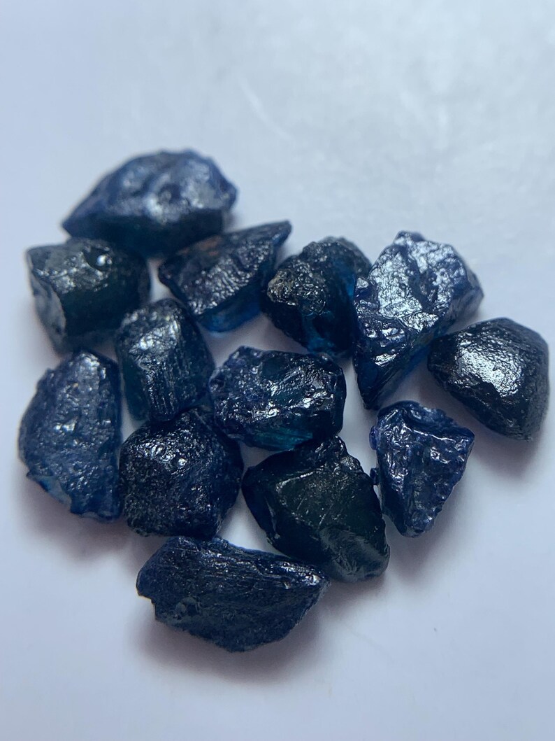 May include: A collection of rough, dark blue gemstones with a textured surface. The stones vary in size and shape, with a deep, saturated color. The gemstones are scattered on a white surface, creating a contrast that highlights their color and texture.
