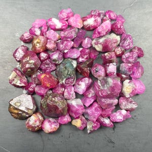Peut inclure: Une collection de rubis bruts et non polis dans diverses nuances de rose vif et de rouge profond. Les pierres présentent des formes irrégulières et dentelées avec des textures cristallines naturelles, certaines incluant des minéraux blancs ou sombres.