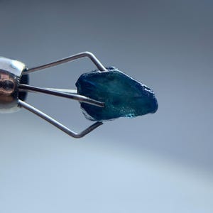 May include: A close-up of a rough, teal-colored gemstone held by a silver metal tool. The gemstone has a textured surface and is illuminated against a soft, blurred background. The tool's design allows for a clear view of the stone.