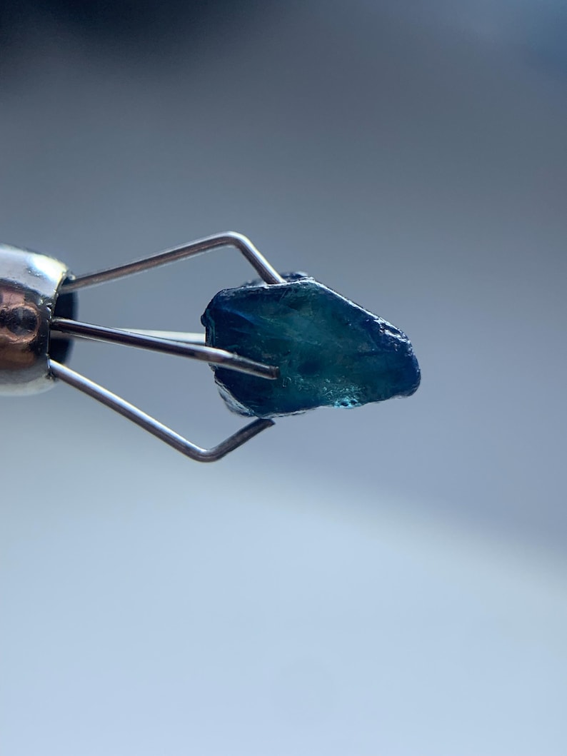May include: A close-up of a rough, teal-colored gemstone held by a silver metal tool. The gemstone has a textured surface and is illuminated against a soft, blurred background. The tool's design allows for a clear view of the stone.