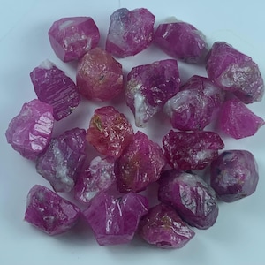 Puede incluir: Una colección de piedras preciosas de rubí en bruto y sin pulir en varios tonos de rosa intenso y magenta. Cada piedra presenta una forma irregular y dentada con texturas cristalinas naturales, bordes translúcidos y ocasionales inclusiones minerales blancas.