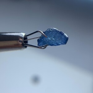 May include: A small, rough-cut, blue gemstone is held by a pair of metal tweezers. The gemstone has a crystalline appearance and is a deep shade of blue. The tweezers are silver-colored and appear to be used for handling delicate items.