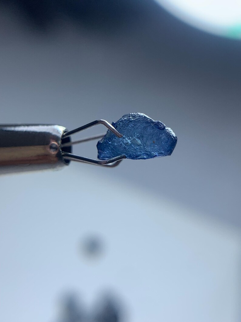 May include: A small, rough-cut, blue gemstone is held by a pair of metal tweezers. The gemstone has a crystalline appearance and is a deep shade of blue. The tweezers are silver-colored and appear to be used for handling delicate items.