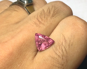 May include: A faceted, triangular pink gemstone. The gem is held in a hand, with a silver ring with a dark blue stone visible in the background. The gemstone's facets reflect light, creating a sparkling effect.