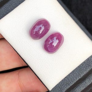 May include: Two polished, oval-shaped, deep red gemstones. The gems are displayed on a white, textured surface within a black box. The stones have a glossy finish, reflecting light.