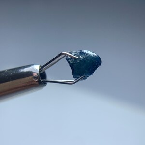 May include: A small, rough-cut, dark blue gemstone is held by a pair of silver tweezers. The gemstone has an irregular shape and a slightly textured surface. The background is a soft, out-of-focus blue.