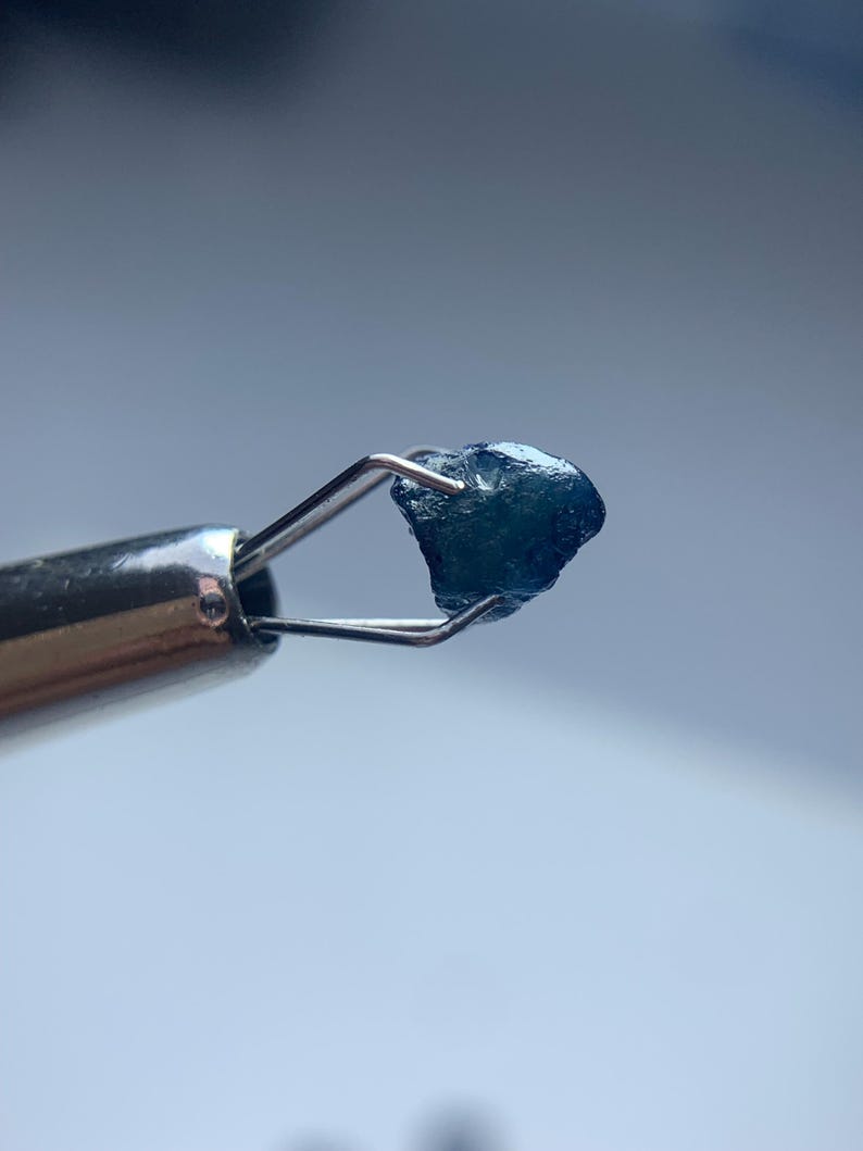 May include: A small, rough-cut, dark blue gemstone is held by a pair of silver tweezers. The gemstone has an irregular shape and a slightly textured surface. The background is a soft, out-of-focus blue.