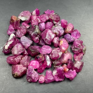 Puede incluir: Una colección de piedras preciosas de rubí en bruto y sin pulir en varios tonos de rosa intenso y magenta. Estos especímenes minerales naturales presentan formas irregulares y dentadas con texturas de translúcidas a opacas con inclusiones blancas.