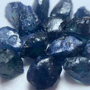 May include: A collection of raw, uncut sapphire gemstones. The sapphires vary in size and shape, displaying a range of deep blue hues with hints of teal and black. The rough texture and natural forms of the stones are visible.