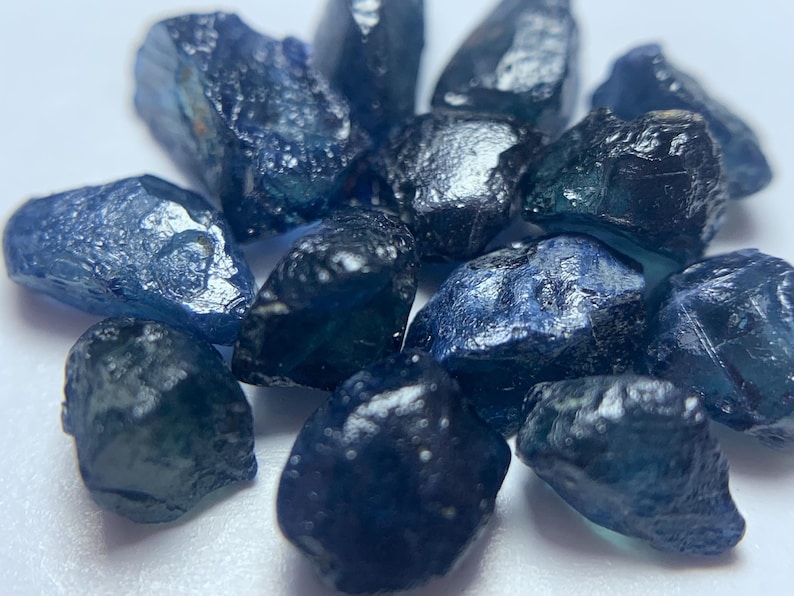 May include: A collection of raw, uncut sapphire gemstones. The sapphires vary in size and shape, displaying a range of deep blue hues with hints of teal and black. The rough texture and natural forms of the stones are visible.