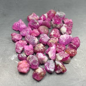 May include: A collection of raw, unpolished ruby gemstones in varying shades of vibrant pink and deep red. These natural mineral stones feature irregular, jagged shapes with translucent to opaque textures and visible white inclusions throughout.