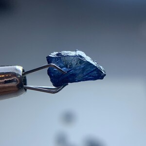 May include: A close-up of a rough, dark blue gemstone held by a pair of silver tweezers. The gemstone has a textured surface and irregular shape, with visible facets. The background is a soft, blurred gradient of blue and white.