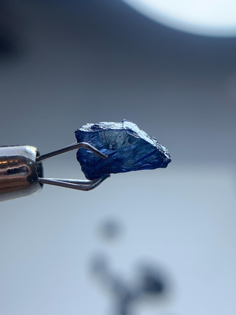 May include: A close-up of a rough, dark blue gemstone held by a pair of silver tweezers. The gemstone has a textured surface and irregular shape, with visible facets. The background is a soft, blurred gradient of blue and white.