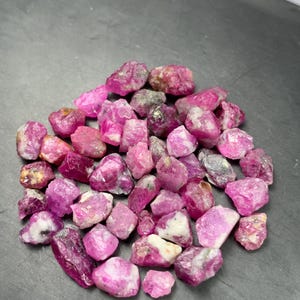 Puede incluir: Una colección de rubíes en bruto y sin pulir en varios tonos de rosa vibrante y magenta intenso. Estos especímenes minerales naturales presentan formas irregulares y dentadas con inclusiones blancas y grises visibles.