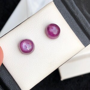 Puede incluir: Dos gemas redondas y pulidas, de color magenta intenso, posiblemente rubíes, se exhiben sobre una superficie blanca. Las gemas tienen una superficie lisa y reflectante, sobre un fondo blanco y negro.