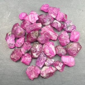 May include: A collection of raw, unpolished ruby gemstones in various shades of vibrant pink and deep magenta. The stones feature irregular, jagged shapes with natural crystalline textures, some showing white mineral inclusions and dark metallic flecks.