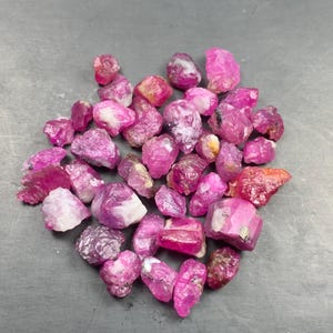 May include: A collection of raw, unpolished ruby gemstones in various shades of vibrant pink and deep magenta. These natural mineral specimens feature irregular, jagged shapes with translucent to opaque textures and visible crystalline structures.