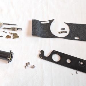 May include: A collection of disassembled camera parts, including a black textured grip, a black metal plate, and various screws and mechanical components. The parts are laid out on a white surface, possibly for repair.