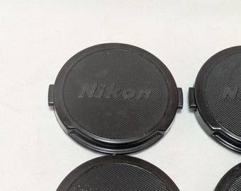 May include: Several black Nikon lens caps are arranged on a white surface. The caps are round with a textured surface and the Nikon logo embossed in the center. Each cap has a small tab on either side.