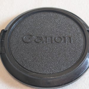 以下が含まれることがあります： 表面に質感のある黒いプラスチック製の円形カメラレンズキャップ。中央にはCanonの文字が浮き彫りになっています。キャップの両サイドには、カメラレンズの前面に固定するためのピンチ式リリースレバーが付いています。
