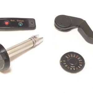 Puede incluir: Cuatro piezas negras de cámara sobre un fondo blanco. Incluye un dial con marcas ASA, una palanca, un panel de botones con el texto "Bat. Check" y un componente plateado y negro.