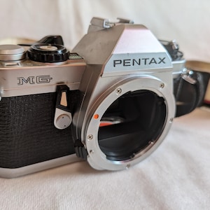 以下が含まれることがあります： レンズマウントと黒い革製グリップを備えた、シルバーとブラックのペンタックスMG 35mmフィルムカメラ。カメラの上部には「PENTAX」、前面には「MG」の文字がプリントされています。カメラストラップが取り付けられています。
