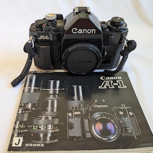 以下が含まれることがあります： 黒いCanon A-1 35mmフィルムカメラ。レンズキャップと黒いストラップが付いています。カメラの下には、Canon A-1の文字と様々なレンズやカメラ本体の画像が印刷された黒い取扱説明書が置かれています。