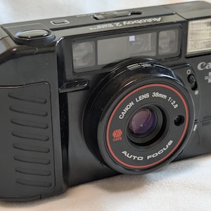 Puede incluir: Cámara de fotos Canon Autoboy 2 Quartz negra de 35 mm. La cámara tiene flash incorporado y una lente Canon de 38 mm de distancia focal y una apertura de f/2.8. La lente tiene las palabras "AUTO FOCUS" y "CAFE".