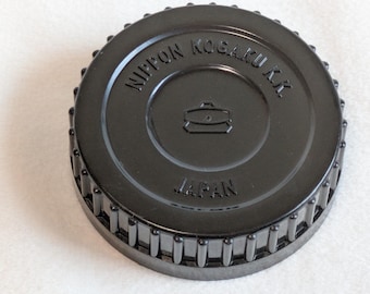 May include: Black camera lens cap with the text "NIPPON KOGAKU K.K." and "JAPAN" in white lettering. The cap has a ridged edge and a central emblem of a camera. A vintage camera accessory.