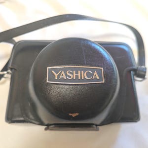 以下が含まれることがあります： 長方形のバッジに銀色の文字でYASHICAと記された、丸みを帯びたレンズカバー付きの黒いレザー製カメラケース。黒いショルダーストラップが付属しており、前面に小さな摩耗が見られます。