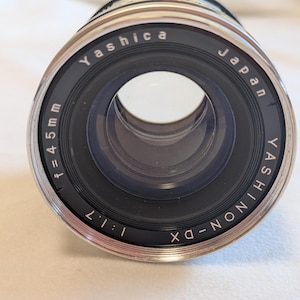 以下が含まれることがあります： 黒とシルバーのデザインのヤシカカメラレンズ。レンズには「Yashica Japan」と「YASHINON-DX」の文字が印刷されています。レンズの焦点距離はf=45mm、絞りは1:1.7です。