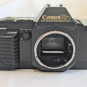 Op de afbeelding: Een zwarte Canon T70 camera met gouden letters. De tekst "Canon T70" is zichtbaar. De camera heeft een zilveren lensvatting en diverse knoppen en draaiknoppen. Een vintage cameramodel, waarschijnlijk uit de jaren 80.