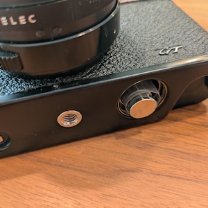 以下が含まれることがあります： 黒いカメラ本体にレンズが付いたクローズアップ。カメラは、ネジマウントと巻き上げノブを含む、テクスチャ加工された表面と銀色のディテールを備えています。レンズは黒色で、「TOPAL ELEC」の文字が入っています。