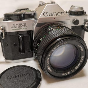 May include: A silver and black Canon AE-1 Program 35mm film camera with a 50mm f/1.4 lens. The lens cap is in front of the camera. The camera has a black strap attached. The text "Canon" and "AE-1 Program" are visible.