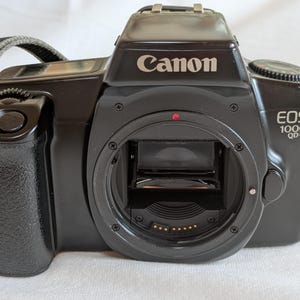 以下が含まれることがあります： 黒いCanon EOS 1000 QD-Pカメラ。レンズマウントとストラップ付き。Canonのロゴとモデル名は白。カメラはテクスチャード加工されたグリップと、レンズマウントの近くに赤い点があります。