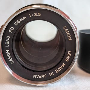 以下が含まれることがあります： 黒いレンズフードが付いた、Canon FD 135mm 1:3.5レンズ。レンズは黒色で、シルバーのアクセントがあり、「CANON LENS FD 135mm 1:3.5 MADE IN JAPAN」の文字が印刷されています。