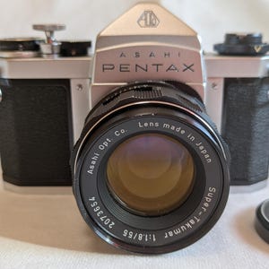 以下が含まれることがあります： Super-Takumar 55mm f/1.8レンズを搭載した、ヴィンテージのAsahi Pentaxカメラ。 カメラ本体はシルバーとブラックで、レンズが際立っています。 レンズキャップが右側にあります。 カメラには「Asahi Pentax」の文字が印刷されています。