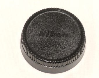 May include: A black Nikon lens cap. The cap is round with a textured surface and a ridged edge. The Nikon logo is embossed in the center of the cap.