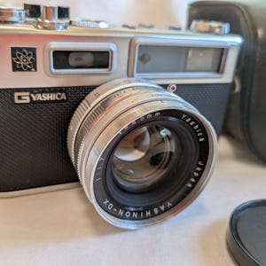 Könnte beinhalten: Silberne und schwarze Yashica Kamera mit einem 45-mm-Objektiv. Die Kamera hat ein schwarzes, strukturiertes Gehäuse und eine silberne Oberseite. Das Objektiv ist mit "Yashica" und "Japan" beschriftet. Eine schwarze Ledertasche ist im Hintergrund.